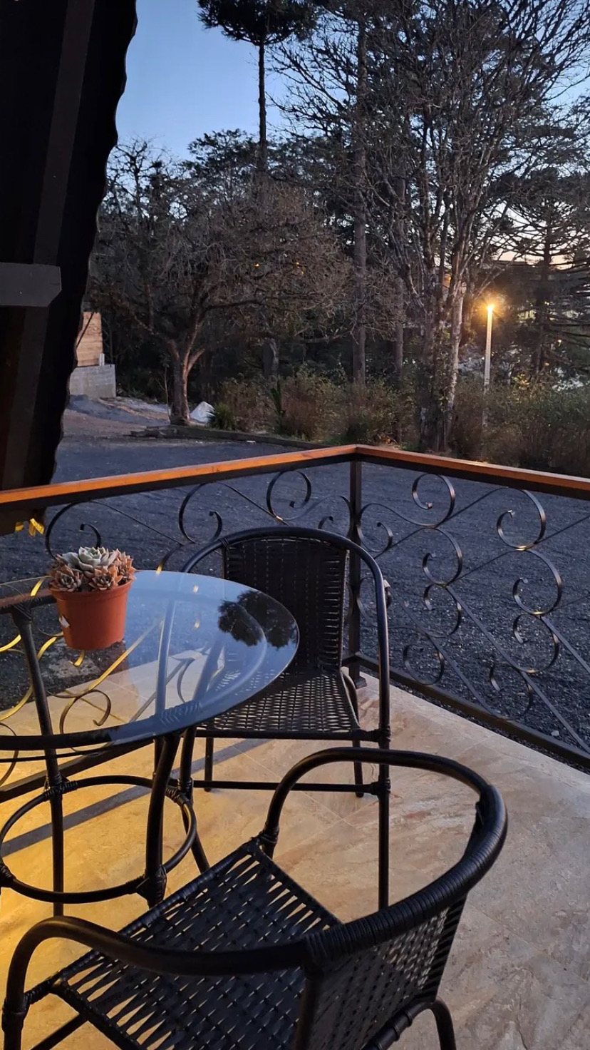Varanda do chalé com mesa e cadeiras de ferro, olhando pras araucárias no fim de tarde